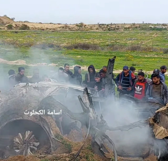 A car that has been hit by an air strike, its almost entirely mangled and there are burn marks and smoke rising out of it. Around a dozen people are standing next to it and looking at the wreckage.