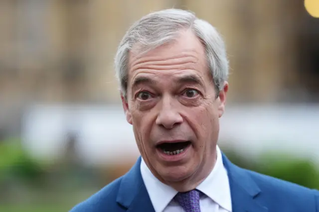 Nigel Farage wearing a blue suit, white shirt and purple tie, with his mouth open and a blurry green and brown background behind him