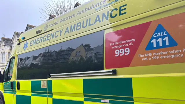 The side of an emergency ambulance, which has a livery of yellow and green alternating panels. On the side there is a sign that reads "For life-threatening emergencies it's 999" and also "Call 111, the NHS number when it's not a 999 emergency"