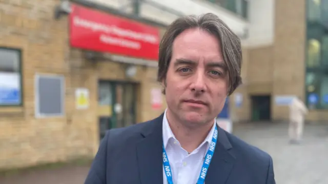 Thom Lafferty looks at the camera - he wears a suit jacket and white shirt, and an NHS lanyard. Blurred behind him is a hospital entrance