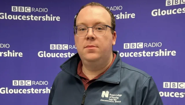 Matthew Stewart looks at the camera - he wears glasses and a blue jacket with a royal college of nursing logo. He is in front of the BBC radio Gloucestershire banner