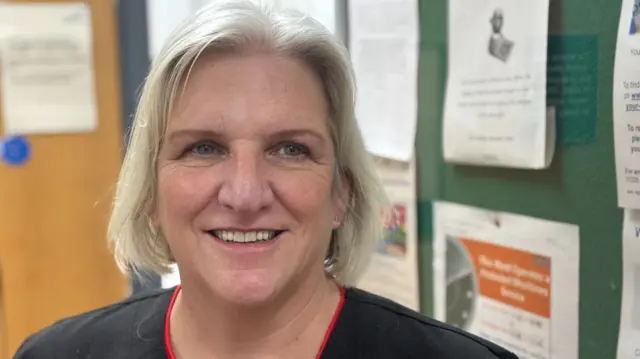 Salli Pilcher smiles at the camera - she wears a black top in the shop, which is of her head and shoulders. Behind her is a noticeboard with various paper pinned to it.