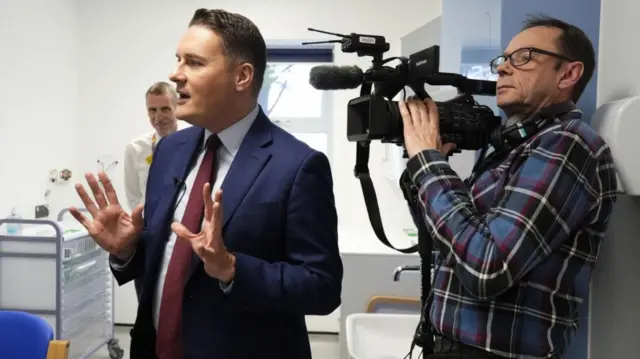 Wes Streeting is pictured with his hands up wearing a blue suit and maroon tie in front of a camerman right behind him