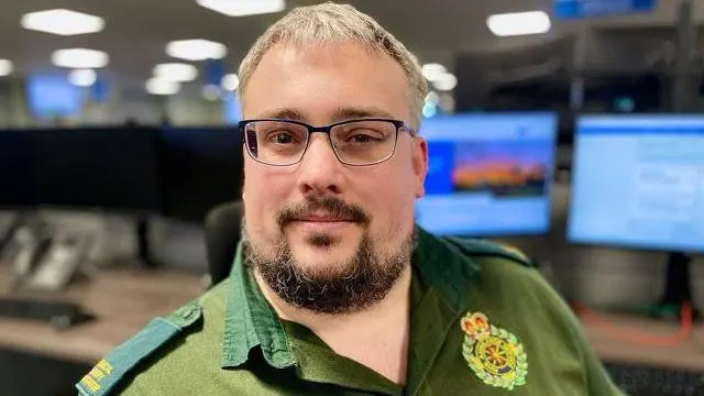 A man with a beard and glasses in green ambulance service uniform sitting in an ambulance 999 call centre with computer screens in the background.