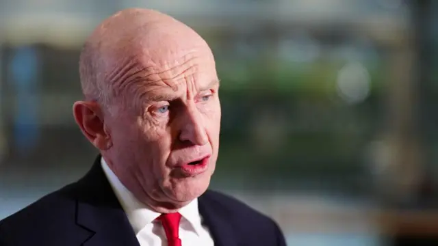 John Healey talking to media, he is wearing a dark suit and a red tie