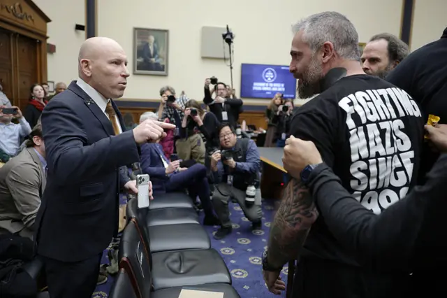 Two men, one bald, one bearded, confront each other in the audience of a congressional hearing