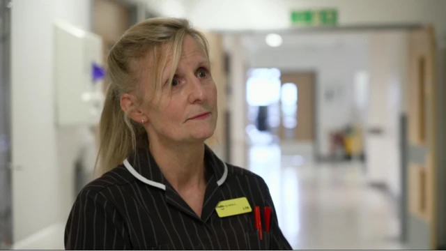 A nurse with blonde hair is looking to the right of the camera, wearing a striped dark uniform with a yellow name badge on. She's standing in the corridor of a hospital ward