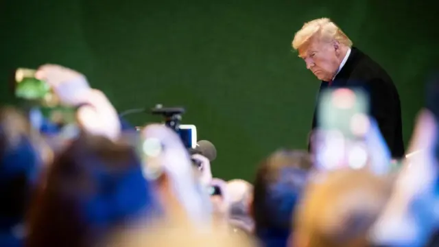 Donald Trump against a green background with blurred out heads of international press in foreground.
