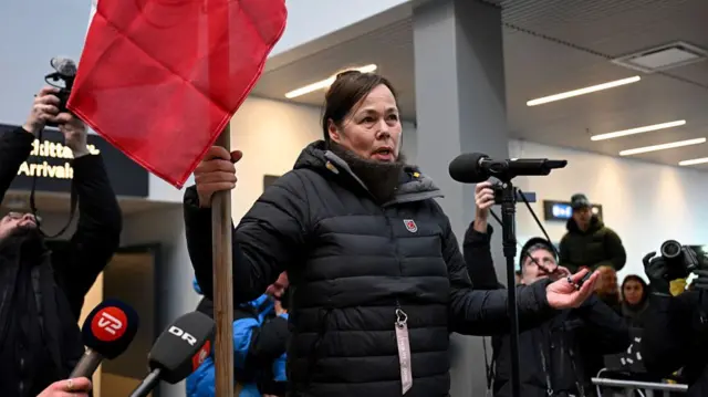 Greenland's Foreign Minister Vivian Motzfeldt holding a Greenland flag as speaks into a microphone. She's wearing wintry layers and a thick coat