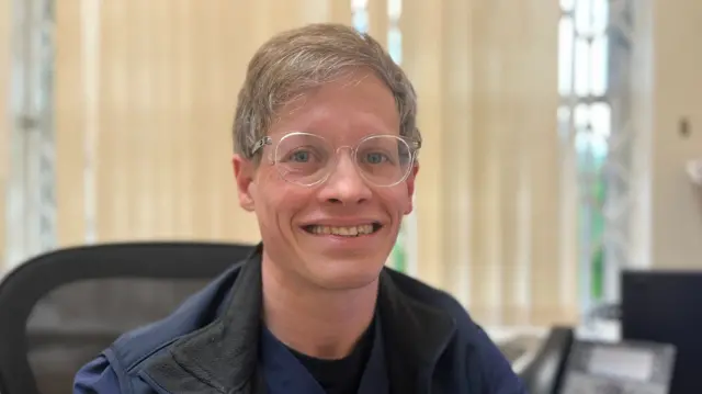 Chris Jacobs smiles at the camera - he wears glasses and a blue top while sitting in a computer chair