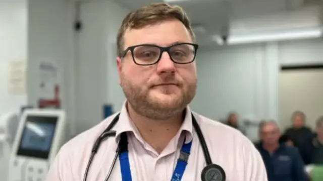 A man wearing glasses, a light pink shirt, a blue lanyard and a stethoscope around his neck looks at the camera