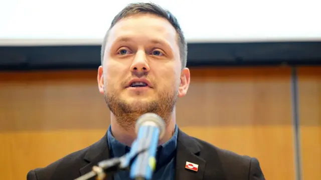 Greenlandic prime minsiter giving a news conference. Jens-Frederik Nielson wears a black blazer with a red and white badge, he is speaking into a microphone