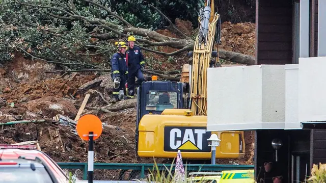 Rescuers standing on soil in front of a yellow excavator