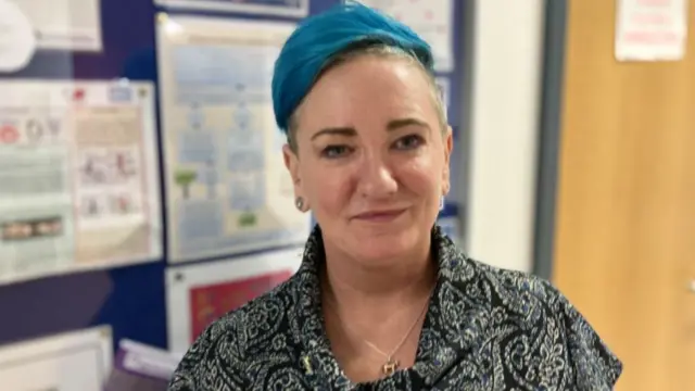 A woman with short blue hair is standing half-smiling in front of a noticeboard