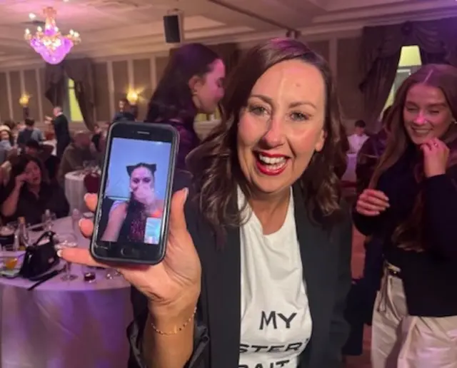 Rachel holds up her phone to camera, it has an image of what appears to be a family member on the other end of a video call