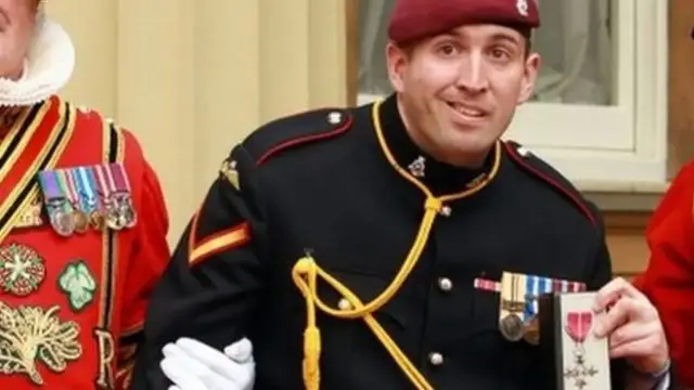 Former British paratrooper Ben Parkinson holding up a MBE medal for his charity work. He is dressed in army uniform and being held up under both arms by men dressed in red suits decorated with medals