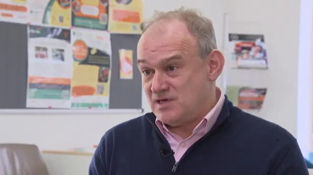 Ed Davey wears a pink shirt and a blue three quarter zip. A school notice board can be seen behind him