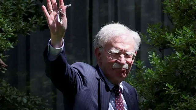 John Bolton wearing a pinstripe jacket, checked red, with clear round glasses and raising his hand in a wave, with car keys in his hand