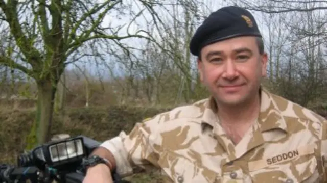 Bob Seddon, former top bomb disposal officer, wearing a brown camouflage jumpsuit with 'Seddon' written on it and a dark blue cap. He has his arm leaning on a piece of equipment and is standing in a woodland