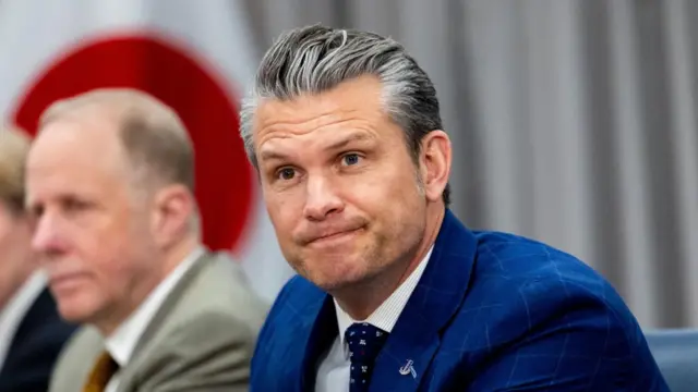Pete Hegseth sat a table in a blue blazer, a Japense flag hangs behind him.