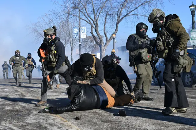 A man being pinned to the floor by an agent. There are several others surrounding him, all hold huge guns and wear gas masks. There's smoke in the air and scenes of chaos