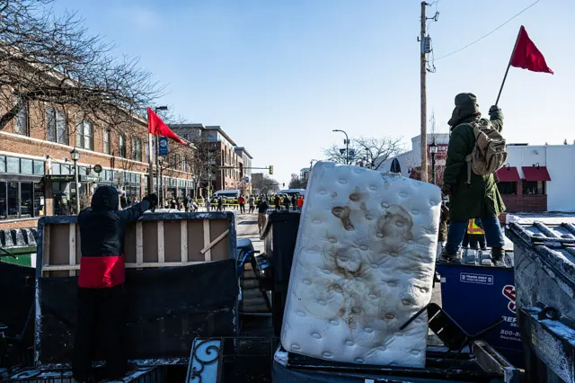 A mattress is being used as a barrier on a street. Two people face away from the camera holding red triangular flags.