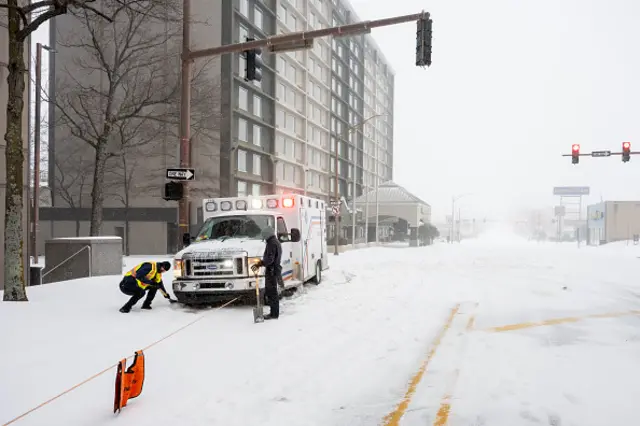 Two workers holding shovels work next to an ambulance on a snowy street