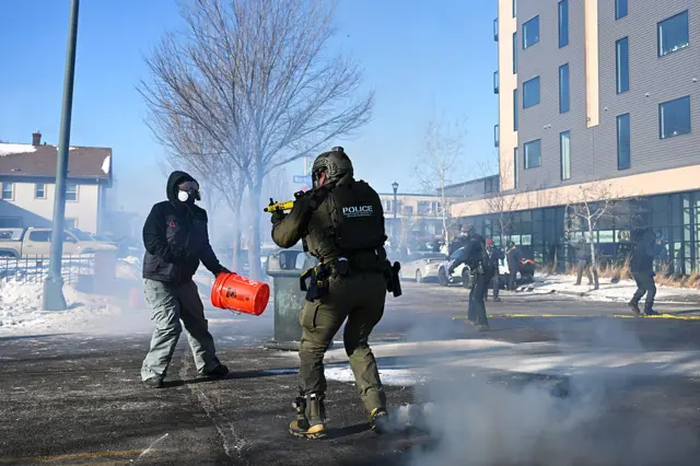 An officer points a gun at a protester at very close range. The protester has one hand in a pocket and the other holding a bucket