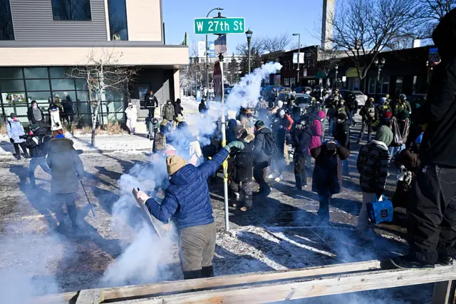 A person throws a smoking canister towards enforcement officers. There's a crowd milling around on a street.