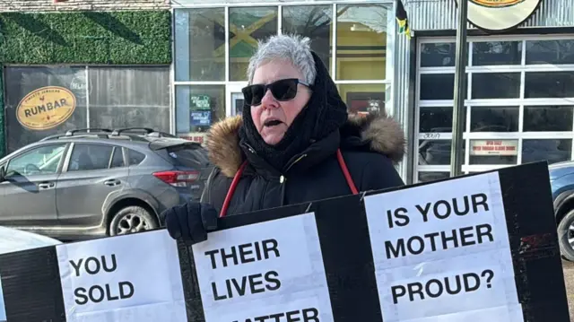 Woman in winter coat and sunglasses holds a protest placard
