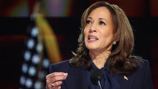 Former US Vice-President Kamala Harris holds her hand up while speaking in front of an American flag.