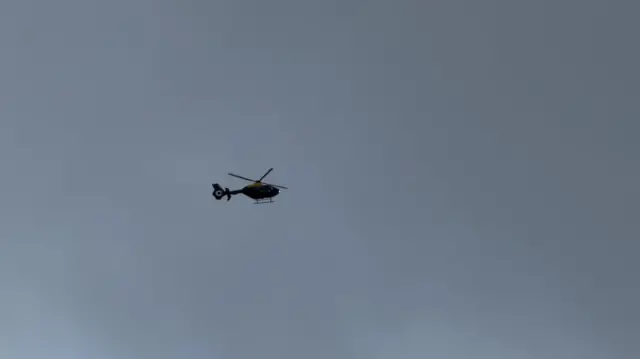 A police helicopter flies high in a grey sky.