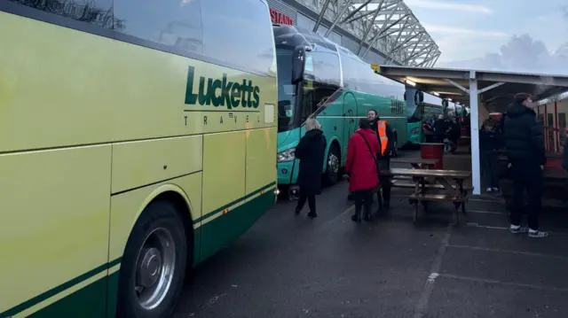 Buses outside a stadium. One is yellow. The other is green.