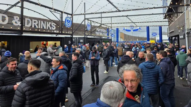 A large crowd of mostly men chat. A few are drinking beer. They are outside a football stadium.