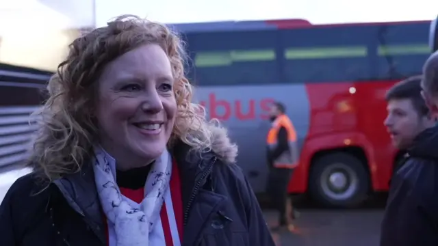 A woman smiles widely. She has blonde curly hair. She looks happy.