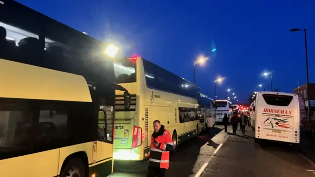 A line of buses in a car park. It is early in the morning.