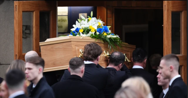 Yorath's coffing being carried into the church by men dressed in black suits. People are gathered outside the entrance doors.