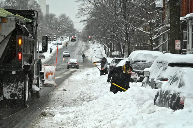 A snow plow heads down Madison Ave. past people shovelling out their snow covered cars during a winter storm
