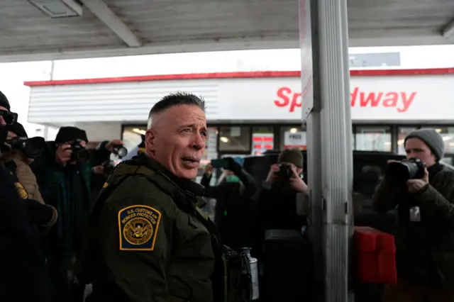 US Border Patrol chief Greg Bovino looks on in a gas station in America. There are journalists with cameras standing around him