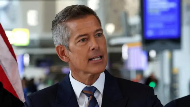 Sean Duffy speaking at an airport. There are information screens and a US flag behind him