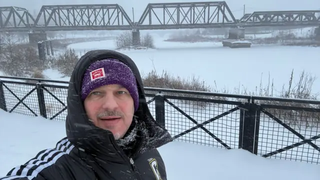 A man in a purple wooly hat with the hood of a black coat pulled over it. A frozen over river is in the background, there is a bridge going over it.