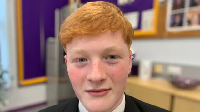 A teenage boy with red hair is looking at the camera. He is wearing a black blazer, white shirt and purple, black and yellow tie.