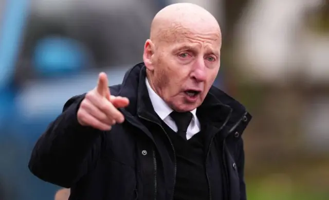 Mickey Thomas outside the church with one finger in the air. Her wears a white shirt, black tie and jumper and a black jacket.