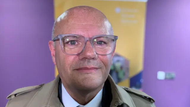 A man with clear rimmed glasses is smiling at the camera. He is standing in front of a purple and yellow background and is wearing a taupe coloured trench coat.