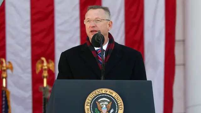Doug Collins stands at a microphone with an American flag behind wearing a black trenchcoat and tie