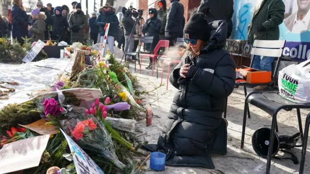 A woman wearing a black jacket and beanie kneels down in front of flowers at a vigil