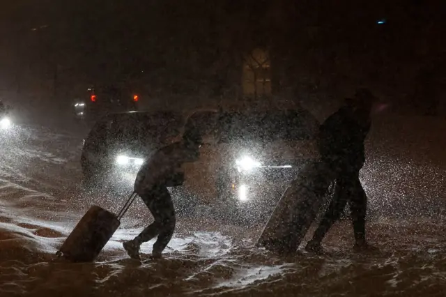 Two people pull heavy suitcases through the snow during a blizzard. There's two cars waiting with their lights on as the two figures cross the street