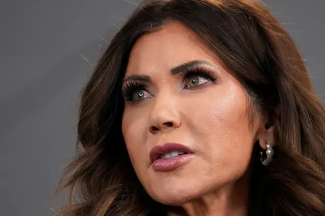 Kristi Noem is close up to the frame. She is looking to the left and may be about to speak . She is wearing pearl earrings and makeup with thick eyelashes and her hair is curled