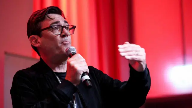 Andy Burnham speaking into a microphone, there is a red background behind him