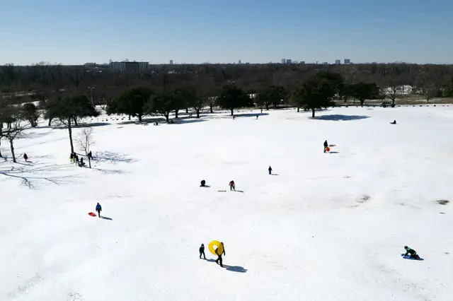 Wide shot of a park in Texas, lawns covered in snow as people walk around with sleighs